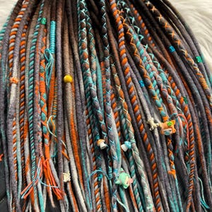 Op de afbeelding: Close-up van een verzameling handgemaakte dreadlocks in verschillende kleuren. De dreads zijn voornamelijk grijs, met accenten van oranje, teal en bruin. Sommige dreads zijn versierd met kralen en decoratieve elementen. Het Dreads Forest-logo is zichtbaar.