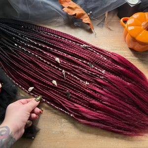 May include: Braided hair extensions in a gradient of black to burgundy. The braids are decorated with silver beads and small charms. A small orange pumpkin-shaped container and a red pumpkin are in the background.