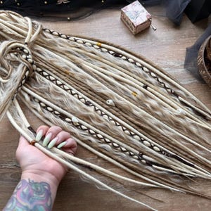 May include: Blonde and brown dreadlocks with decorative accents. The dreadlocks are arranged in a bundle, with some strands held by a hand. The dreadlocks have dark brown and white accents, with small beads and decorative stitching.