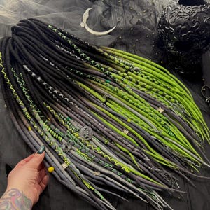 May include: Black, green, and gray synthetic dreadlocks with green and black accents. The dreadlocks are arranged in a fan shape and are lying on a black surface.