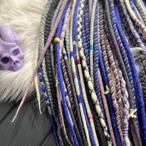 a variety of colorful braided hair accessories, including braids and braided strands, arranged on a wooden surface. There is also a purple skull-shaped object placed next to the hair accessories.