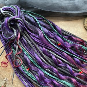 May include: A collection of purple, gray, and teal synthetic dreadlocks. The dreads are adorned with green braids and small cherry charms. The overall style is a blend of colors and textures.