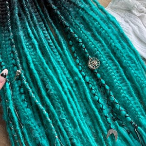 May include: Turquoise and black ombre dreadlocks with silver accents and decorative charms. The dreads are braided with black thread and adorned with a sun and moon charm and a pair of angel wings. The Dread5 Forest logo is visible in the background.