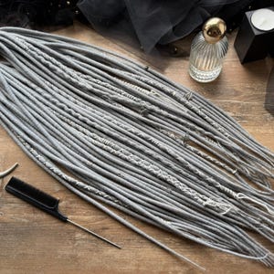 May include: A bundle of gray dreadlocks with white decorative stitching. A black comb and a silver crescent moon pendant are also visible. The dreadlocks are tied together with a yellow band. The Dread5 Forest logo is in the lower left corner.