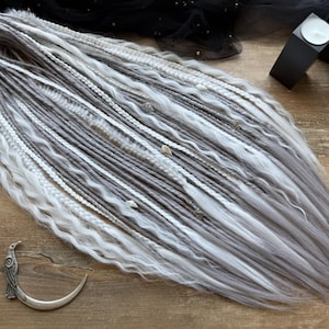 Könnte beinhalten: Ein Set aus grauen und weißen Dreadlocks mit gewellten und geflochtenen Texturen. Die Dreadlocks liegen auf einer Holzoberfläche, mit dekorativen Akzenten und einem halbmondförmigen Haaraccessoire. Das Dreads Forest-Logo ist sichtbar.