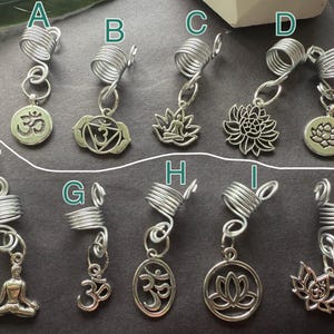 May include: A collection of silver-toned dreadlock beads with various charms. Charms include the Om symbol, lotus flowers, and a seated Buddha. The beads are made of coiled wire and are designed to be inserted into dreadlocks.