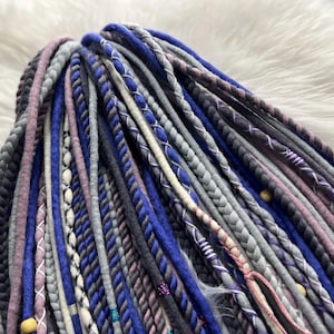 A close-up view of a variety of colorful braided hair accessories, including braided strands in shades of blue, gray, purple, and white, arranged on a white fur surface.