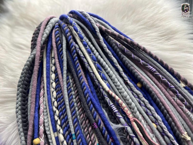 A close-up view of a variety of colorful braided hair accessories, including braided strands in shades of blue, gray, purple, and white, arranged on a white fur surface.