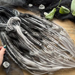 May include: A set of gray and black dreadlocks, with a braided section and silver accents. The dreads are arranged on a wooden surface with a dark gray tulle fabric. The Dread5 Forest logo is visible.
