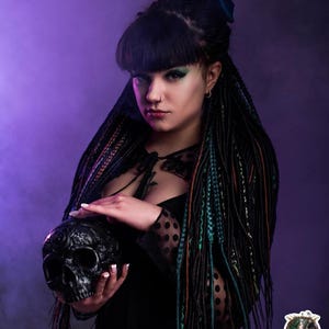 Side view of long synthetic dreads in black and forest green ombre. The model is sitting in a smoky, mystical room with vintage decor. The photo highlights the length of the dreadlock extensions and how they blend with natural dark hair.