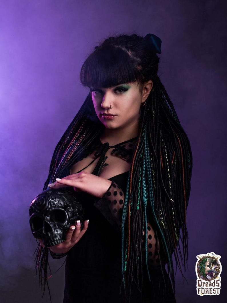 Side view of long synthetic dreads in black and forest green ombre. The model is sitting in a smoky, mystical room with vintage decor. The photo highlights the length of the dreadlock extensions and how they blend with natural dark hair.