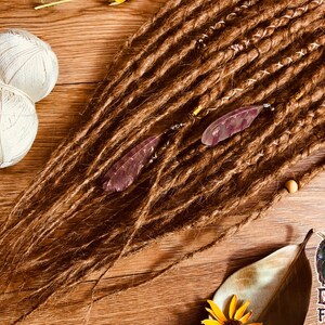 K&ouml;nnte beinhalten: Braune Dreadlocks mit dekorativen Akzenten, darunter kleine Perlen und federf&ouml;rmige Anh&auml;nger. Zwei Kn&auml;uel wei&szlig;er Wolle sind sichtbar, zusammen mit einem Logo mit der Aufschrift "Dread5 FOREST". Die Dreadlocks sind auf einer Holzoberfl&auml;che angeordnet.