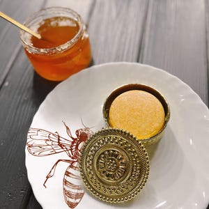 May include: A gold metal container with a round, orange-colored soap bar inside. The container has a decorative lid with a floral design. The container is sitting on a white plate with a bee design.
