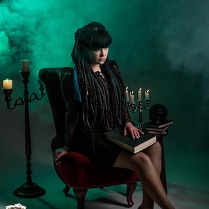 Front view of a model with a full set of forest green ombre dreadlocks and braids. The hair extensions are draped over her shoulders, showing the volume and length. She sits at a mystical table with candles and vintage books, creating a dark boho or viking aesthetic.