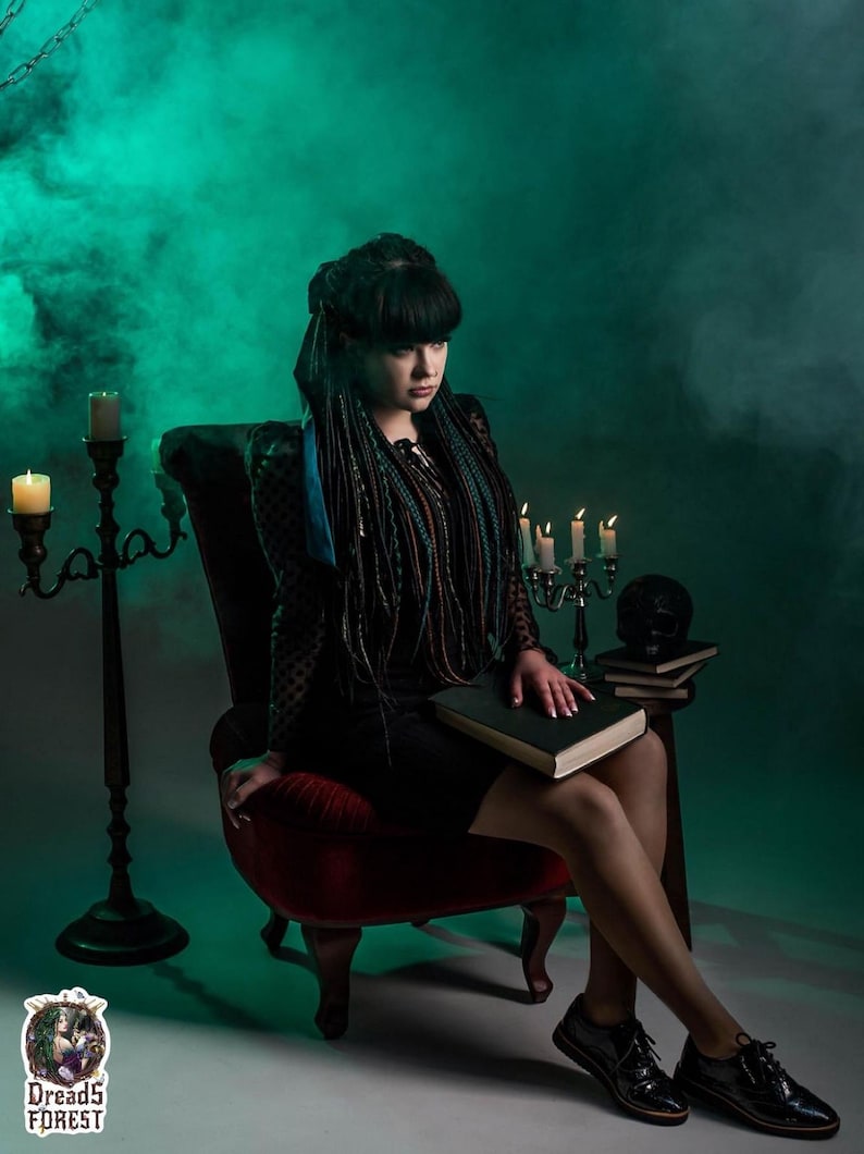 Front view of a model with a full set of forest green ombre dreadlocks and braids. The hair extensions are draped over her shoulders, showing the volume and length. She sits at a mystical table with candles and vintage books, creating a dark boho or viking aesthetic.