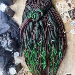 Pode incluir: Um conjunto de extensões de cabelo sintético com uma mistura de fios castanhos escuros e verde-floresta profundo. O cabelo é estilizado numa combinação de tranças apertadas e madeixas onduladas, acentuadas com pequenos berloques prateados.