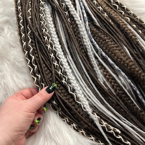 May include: A collection of brown and white synthetic dreadlocks with a variety of textures and styles. The dreadlocks are arranged in a fan shape and are displayed on a white fluffy surface.