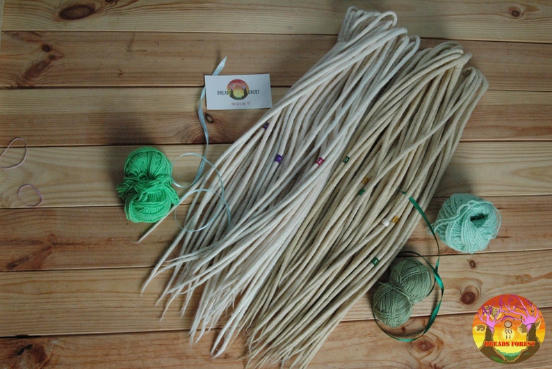 May include: A bundle of white wool dreadlock extensions with green yarn and beads. The dreadlocks are arranged in a fan shape on a wooden surface. The text "Beads Forest" is visible in the lower right corner of the image.