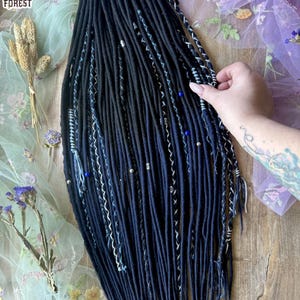Könnte beinhalten: Dunkelblaue Dreadlocks mit weißen und blauen Akzenten, einige Strähnen sind mit Perlen verziert. Die Dreadlocks werden auf einer Holzoberfläche mit floralem Hintergrund präsentiert. Das Logo "Dreads Forest" ist sichtbar.