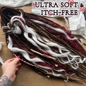 May include: A collection of brown, burgundy, and white dreadlocks with gold accents. The text "ULTRA SOFT & ITCH-FREE" is visible. The Dreads Forest logo is in the upper left corner. A white bird figurine is on the left.