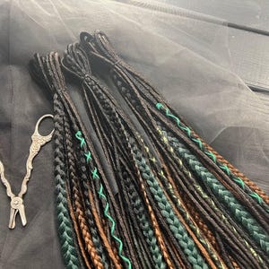 a collection of braided hair accessories, including braids and cords, arranged on a gray surface. There are also a pair of scissors and a green object, possibly a piece of fruit, in the background.