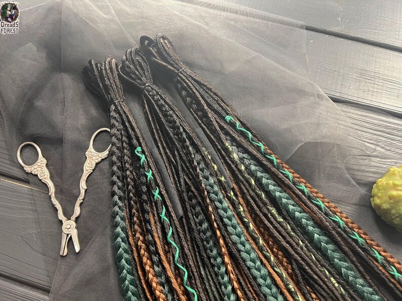 a collection of braided hair accessories, including braids and cords, arranged on a gray surface. There are also a pair of scissors and a green object, possibly a piece of fruit, in the background.