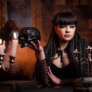 A model sitting in a red velvet vintage chair, showcasing a full set of forest green and black synthetic dreadlocks. She is surrounded by candles, smoke, and mystical decor. The image highlights the natural flow and length of the mixed texture braids and dreads in a lifestyle setting.