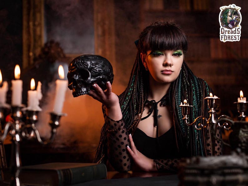 A model sitting in a red velvet vintage chair, showcasing a full set of forest green and black synthetic dreadlocks. She is surrounded by candles, smoke, and mystical decor. The image highlights the natural flow and length of the mixed texture braids and dreads in a lifestyle setting.