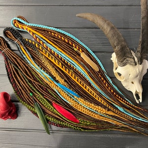 May include: A set of colorful synthetic dreadlocks with brown, yellow, blue, green, and red strands. The dreadlocks are arranged on a wooden surface with a skull and feathers.