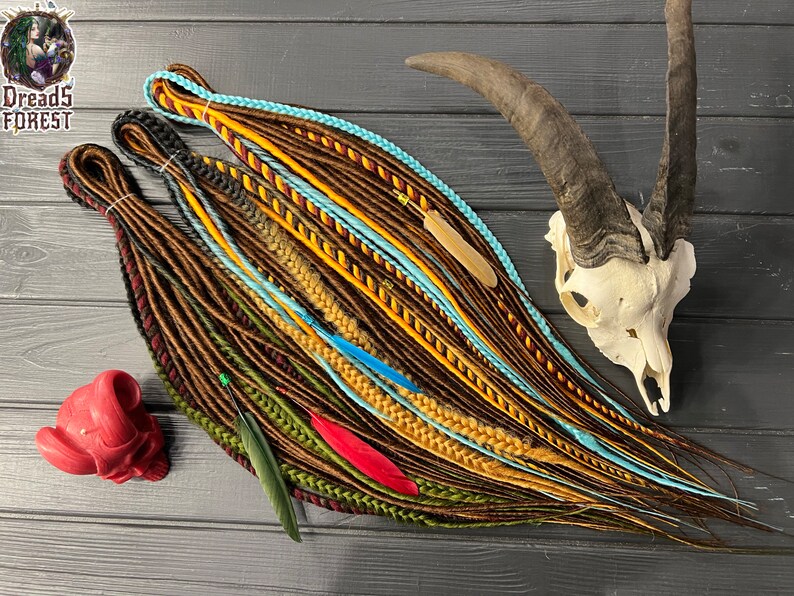 May include: A set of colorful synthetic dreadlocks with brown, yellow, blue, green, and red strands. The dreadlocks are arranged on a wooden surface with a skull and feathers.