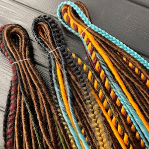 May include: Three sets of synthetic dreadlocks in brown, black, and blue with orange, yellow, and red accents. The dreadlocks are arranged in a row on a dark wooden surface.