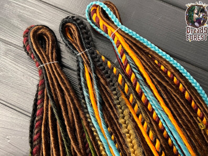 May include: Three sets of synthetic dreadlocks in brown, black, and blue with orange, yellow, and red accents. The dreadlocks are arranged in a row on a dark wooden surface.