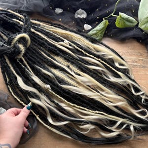 May include: Black and blonde dreadlocks, artfully arranged. The synthetic hair extensions are a mix of black and light blonde, with a knotted base. The image includes a logo that says "Dreads Forest".