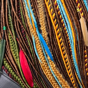 May include: A collection of brown, green, blue, and yellow synthetic dreadlocks with feathers. The dreadlocks are arranged in a fan shape and are tied together with beads.
