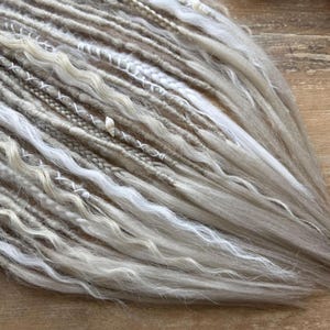 a close-up view of a collection of variously braided and woven hair strands, with a mix of white, gray, and brown colors. The strands appear to be made of yarn or similar materials, and they are arranged in a way that creates a textured and intricate appearance.