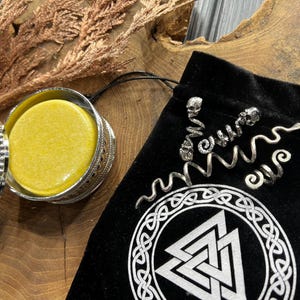May include: A round, yellow soap bar in a silver container with an ornate design. The container is open and the soap is visible. The container is sitting on a wooden surface. A black velvet bag with a silver design is in the background.