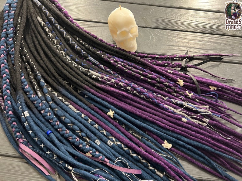 Puede incluir: Un conjunto de rastas sint&eacute;ticas en negro, azul oscuro y morado con detalles blancos y grises. Las rastas est&aacute;n atadas con cintas moradas y negras.
