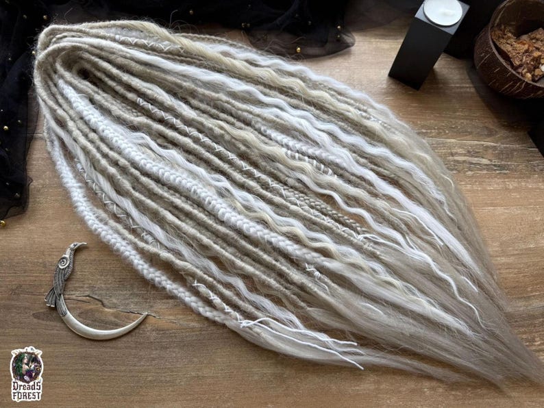 a collection of long, white braided hair strands arranged on a wooden surface. There is also a small metal object, possibly a hair clip, placed next to the hair strands.