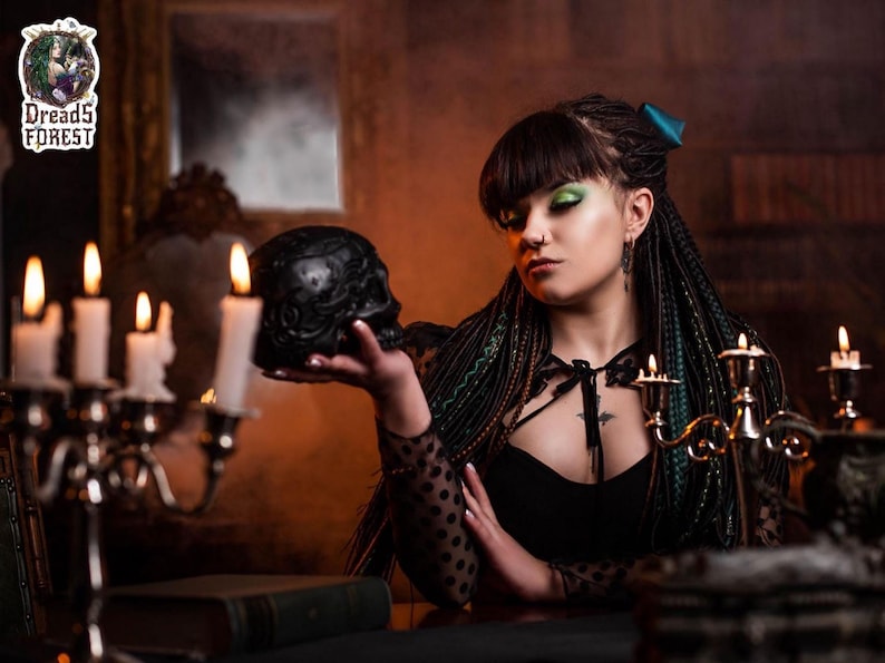 Back view of a full set of forest green and black synthetic dreadlocks and braids. The hair is tied with a large teal satin bow, showing how the extensions look from behind. The photo highlights the mix of textures and the natural fall of the long dreads against a dark green smoky background.