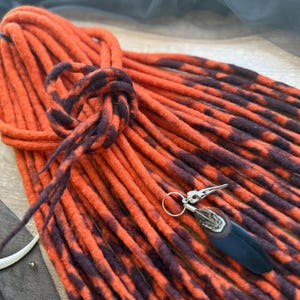 May include: Orange and dark brown dreadlocks with a decorative charm. The dreadlocks are tied in a knot, and the charm features a bird skull design. The dreadlocks are a vibrant orange with dark brown streaks.