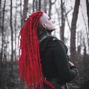 Pode incluir: Uma pessoa com dreadlocks vermelhos e a cabeça raspada está vestindo um casaco preto com capuz e um cinto de couro preto com ferragens prateadas. A pessoa está em pé em uma floresta com árvores nuas.