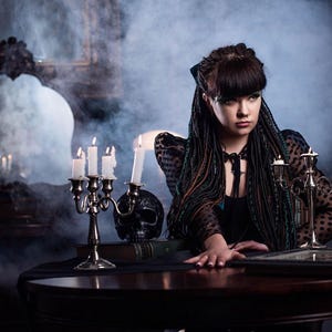 A full-body shot of a model wearing a set of green and black ombre synthetic dreadlocks. She is dressed in a black gothic outfit, holding a book and scissors amidst hanging chains. This photo demonstrates how the long dreadlock extensions look when worn, reaching below the waist.