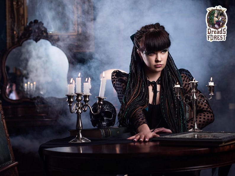 A full-body shot of a model wearing a set of green and black ombre synthetic dreadlocks. She is dressed in a black gothic outfit, holding a book and scissors amidst hanging chains. This photo demonstrates how the long dreadlock extensions look when worn, reaching below the waist.