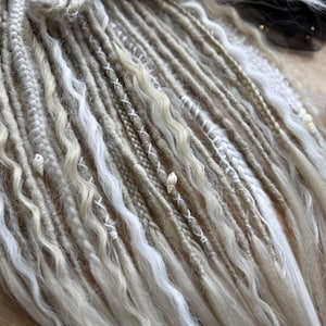 A close-up view of various braided and twisted hair strands in different shades of white and beige, arranged on a wooden surface.