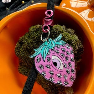 Op de afbeelding: Een roze aardbei-bedel met meerdere oogontwerpen en een turquoise groene stengel en bladeren. De bedel is bevestigd aan een roze draadwikkel en een zwarte dreadlock. De bedel zit in een oranje kom.
