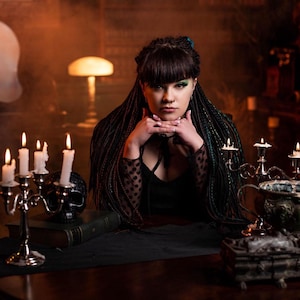 A close-up portrait of a model with green and black ombre synthetic dreadlocks and intricate fishtail braids. She is holding a black skull against a purple smoky background. The lighting emphasizes the mixed textures and thin diameter of the handmade dreadlock extensions.