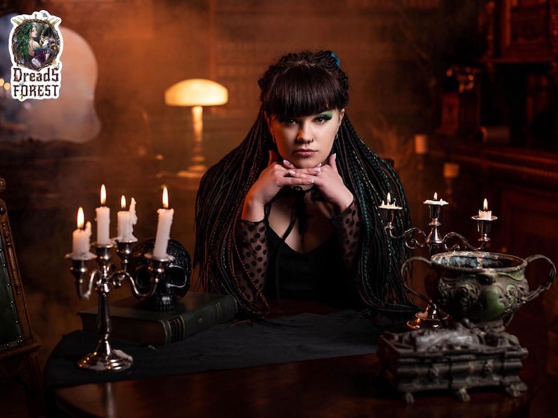 A close-up portrait of a model with green and black ombre synthetic dreadlocks and intricate fishtail braids. She is holding a black skull against a purple smoky background. The lighting emphasizes the mixed textures and thin diameter of the handmade dreadlock extensions.