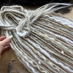 A hand holding a bundle of long, white braided hair with various decorative elements attached to it, such as beads and sequins.