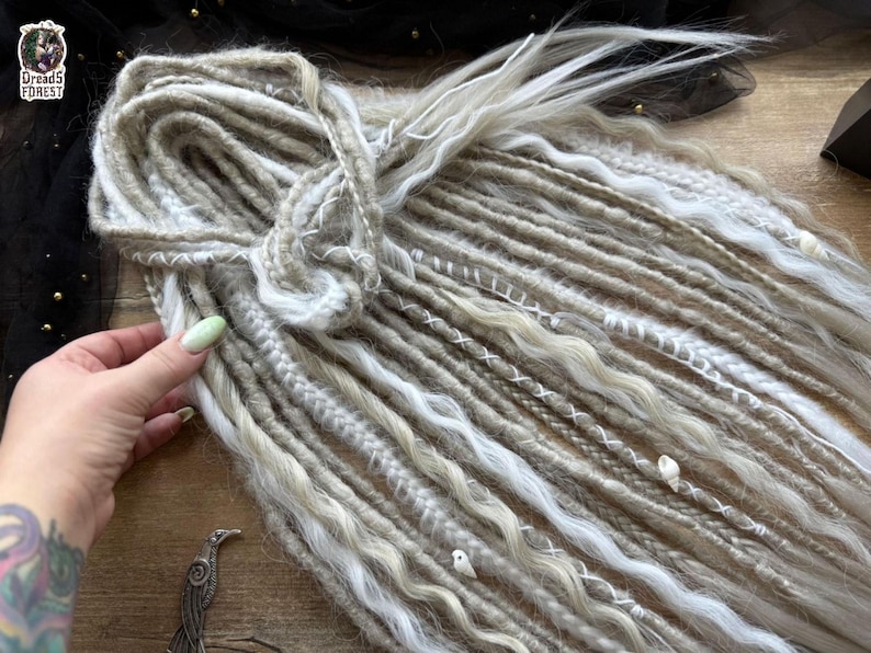 A hand holding a bundle of long, white braided hair with various decorative elements attached to it, such as beads and sequins.