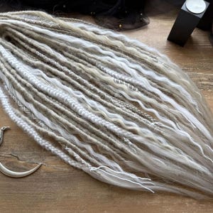 a collection of long, white braided hair strands arranged on a wooden surface. There is also a small metal object, possibly a hair clip, placed next to the hair strands.